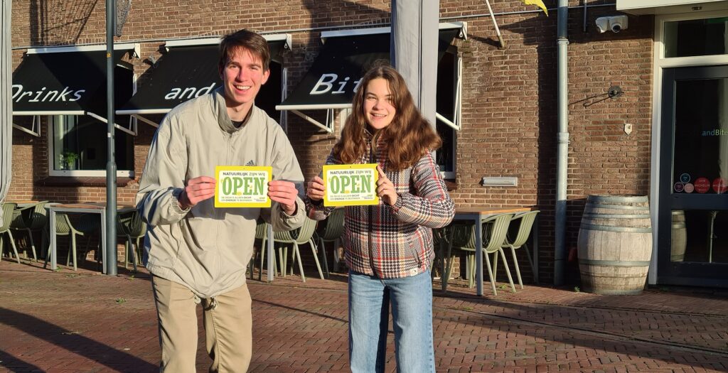Man en vrouw houden bordjes vast met de tekst: Natuurlijk zijn wij open. De deur is alleen dicht om energie te besparen.